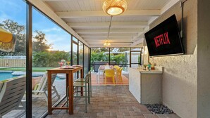 A covered outdoor patio with a pool, a dining area with chairs and a table, and a TV mounted on the wall. - Backyard Paradise • Sleeps 14 • Arcade Games • Spa (Bradenton)