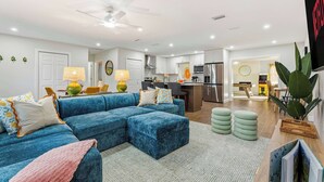A modern living room with a blue sectional sofa, a ceiling fan, and a kitchen in the background. - Backyard Paradise • Sleeps 14 • Arcade Games • Spa (Bradenton)