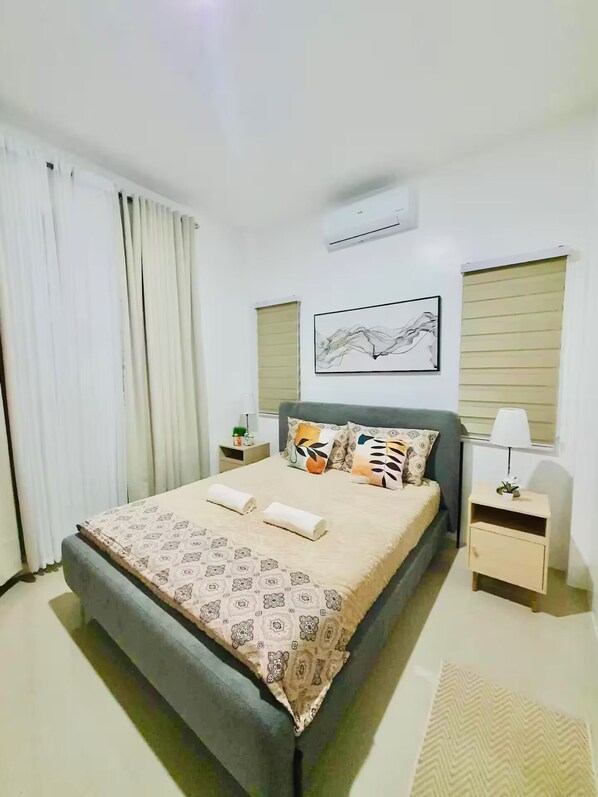 A bedroom with a bed, two bedside tables, a nightstand, a window with blinds, and a wall-mounted air conditioner.
