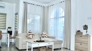 Un salon moderne avec un canapé, une table basse et une petite table d’appoint. On y trouve une fenêtre avec des rideaux légers, un radiateur et une étagère murale agrémentée d’objets décoratifs.