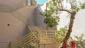 Un escalier avec des rampes jaunes mène à un bâtiment. On y voit un arbre aux feuilles vertes et quelques plantes en pot.
