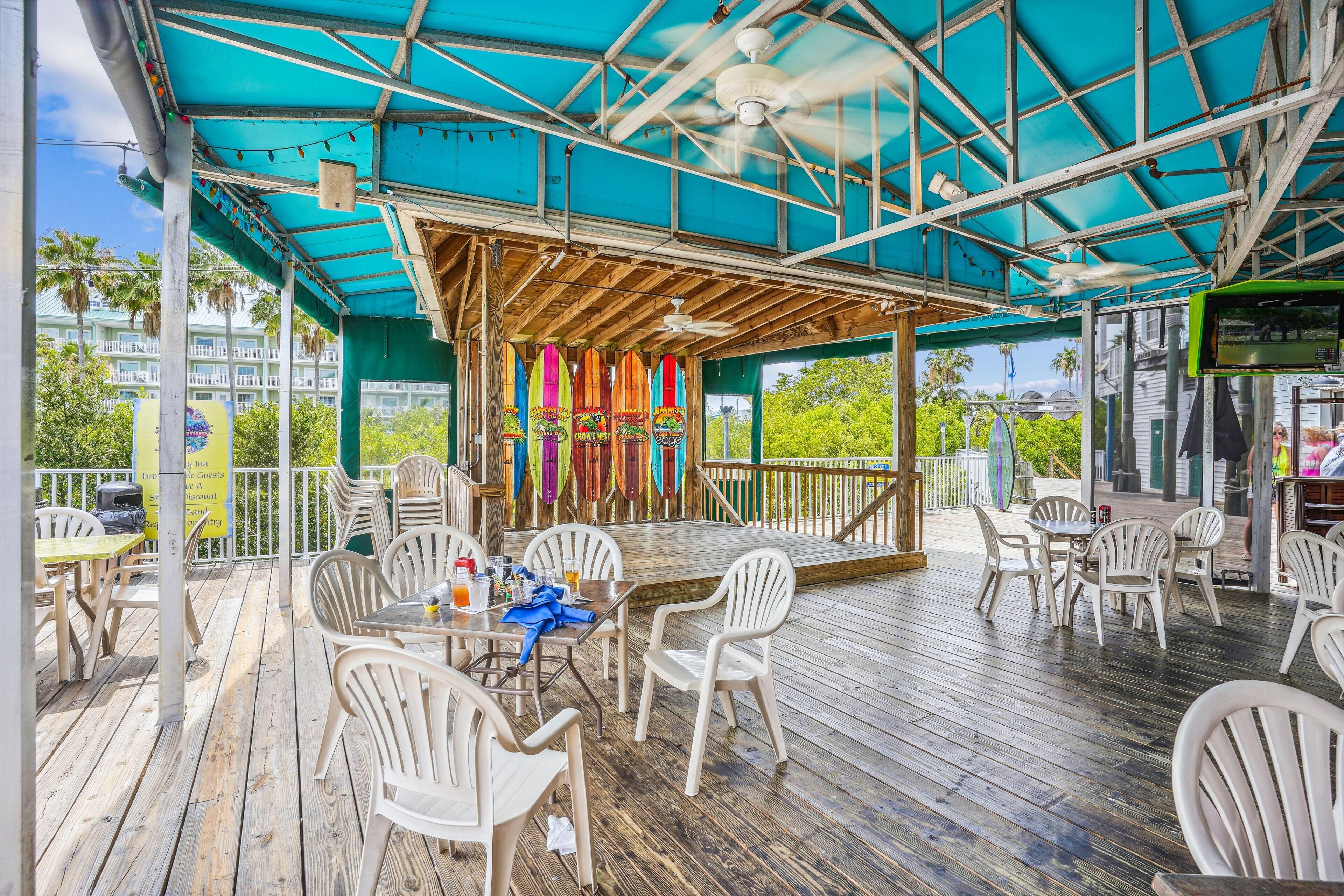 Een terras met houten tafels en stoelen, een overdekt zitgedeelte met kleurrijke decoraties, en een uitzicht op palmbomen en gebouwen op de achtergrond.