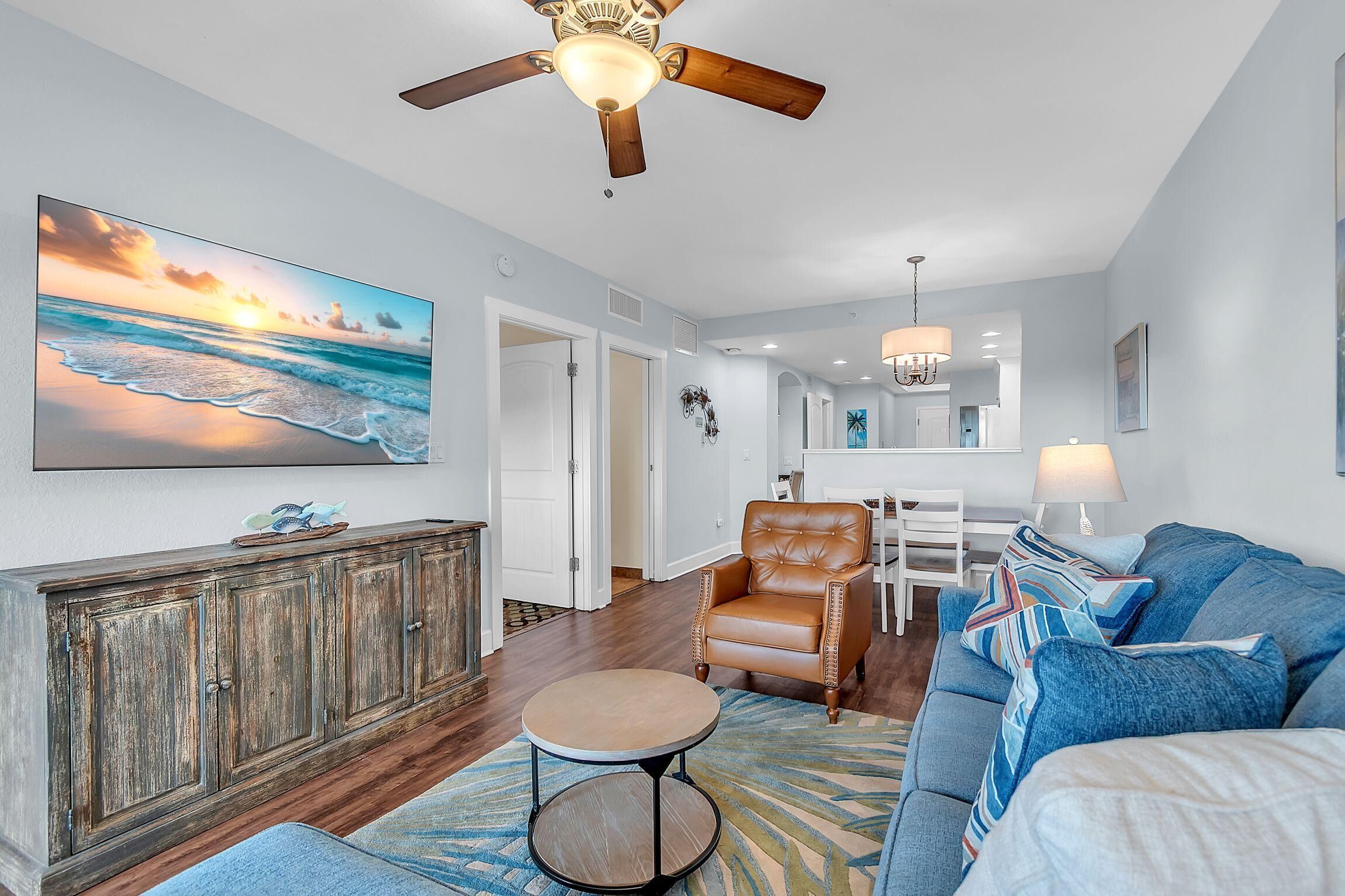 A spacious living room with a large ocean view painting, a leather armchair, a round coffee table, and a ceiling fan.