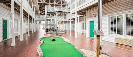 A spacious indoor area with a green artificial turf mini-golf course, white wooden railings, and a high ceiling with exposed beams.