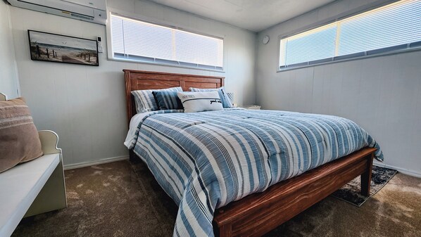 Un dormitorio con una cama de madera, ropa de cama a rayas, una mesita de noche blanca, una silla marrón y un cuadro en la pared.