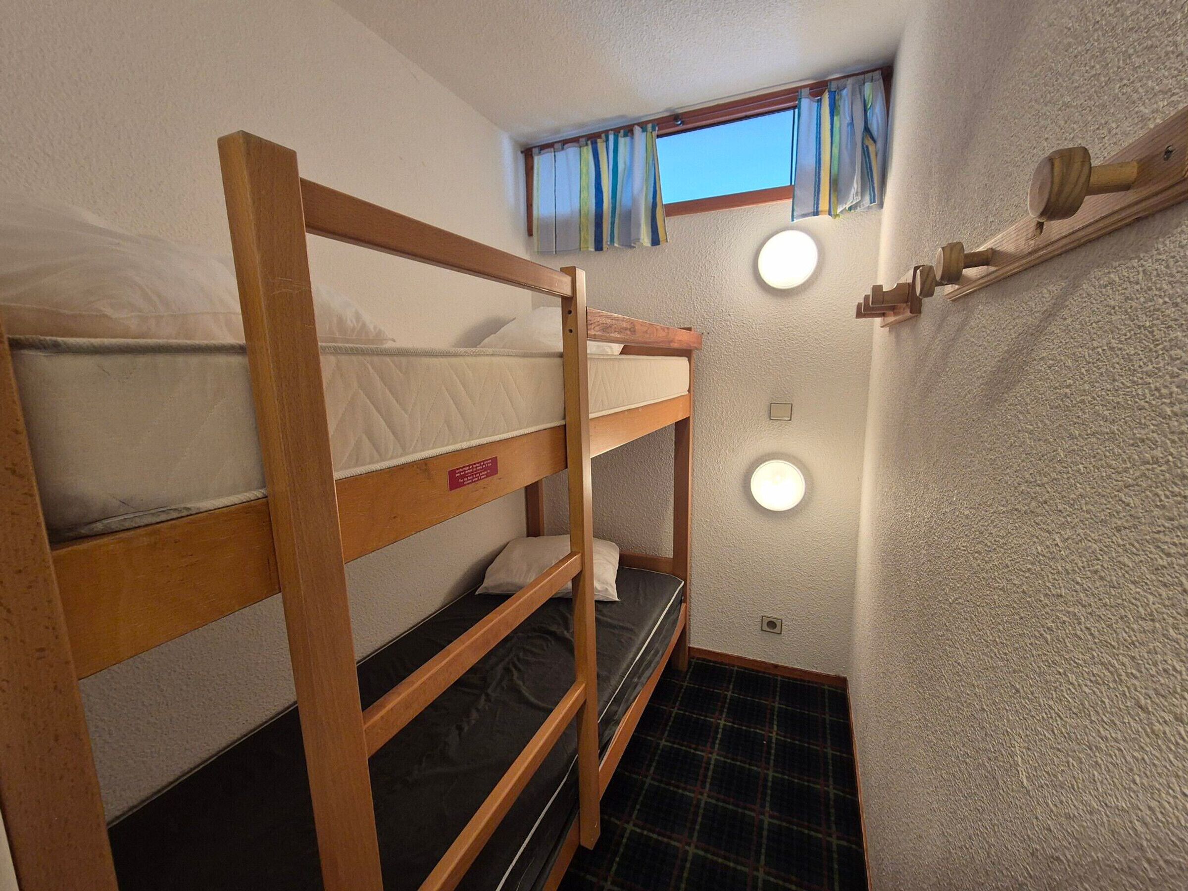 Une chambre avec un lit superposé, une fenêtre, deux lampes fixées au mur et une petite étagère sur laquelle se trouve un objet en bois.