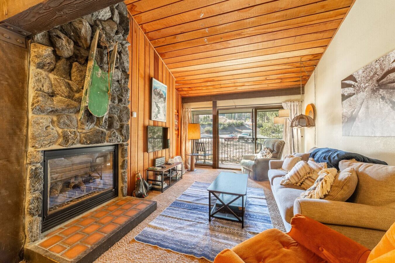 A cozy living room with a stone fireplace, wooden ceiling, and a view of the outdoors.