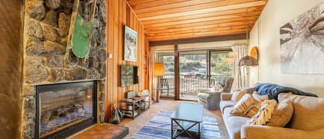 A cozy living room with a stone fireplace, wooden ceiling, and a view of the outdoors.