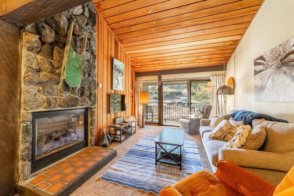 A cozy living room with a stone fireplace, wooden ceiling, and a view of the outdoors.