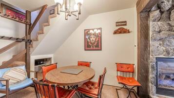 A cozy room with a round wooden table, orange leather chairs, a chandelier, a staircase with a 'Welcome' sign, and a stone fireplace.