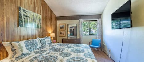 A bedroom with a large bed, a wooden wall, a window with curtains, a chair, and a TV mounted on the wall.