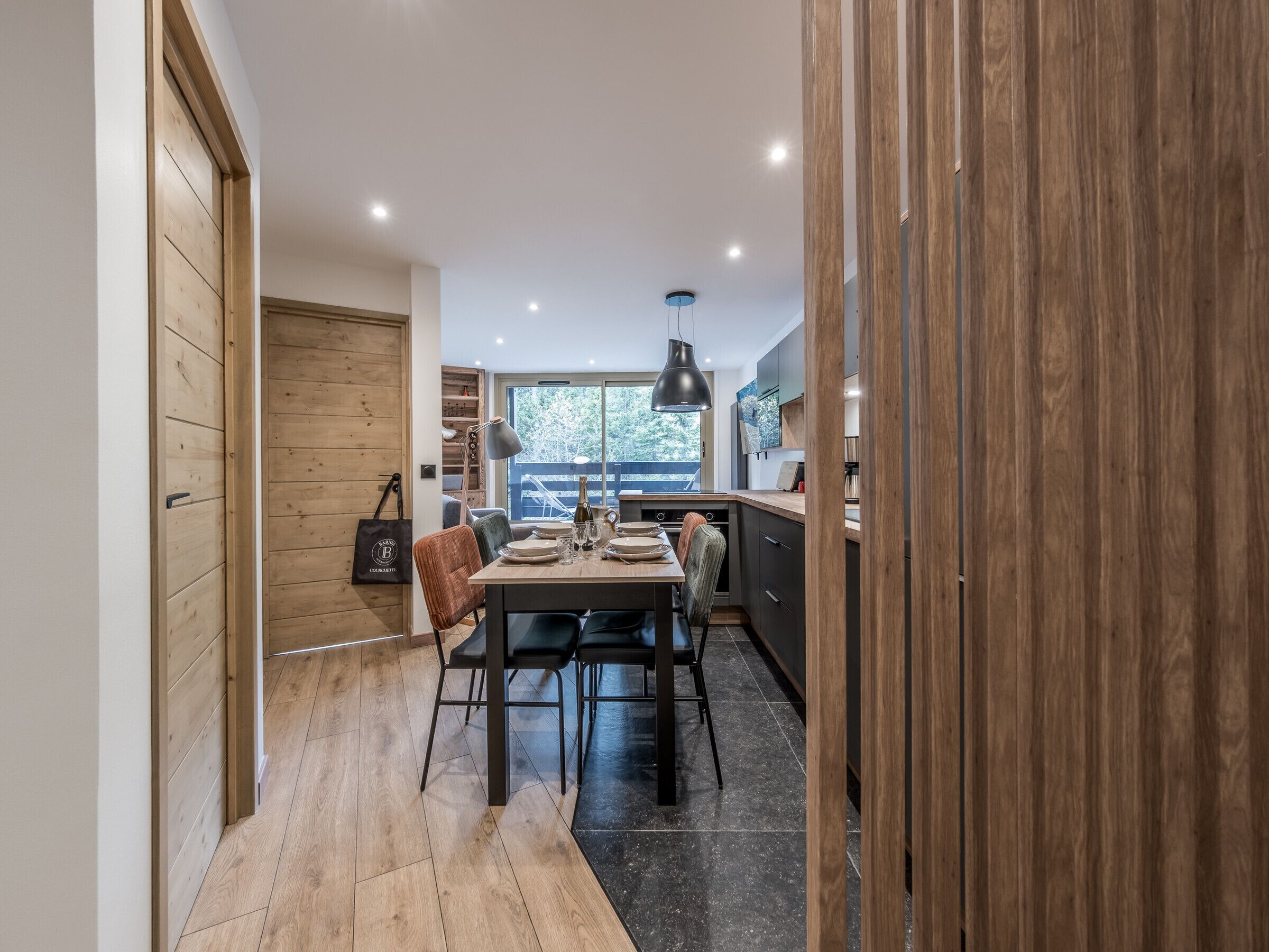 Une cuisine moderne avec des meubles en bois, un coin repas avec une table dressée pour quatre personnes, et une vue sur l’extérieur grâce à une grande fenêtre.