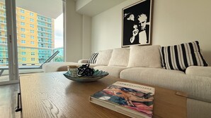 Un salon moderne avec un canapé blanc, une table basse sur laquelle se trouvent un livre et un bol, et une grande fenêtre donnant sur un immeuble jaune.