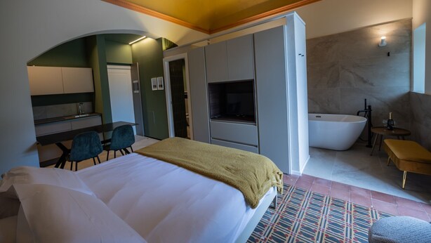 Quarto de hotel com cama, escrivaninha, cadeira, televisão, banheira e uma pequena mesa com garrafa e copo.