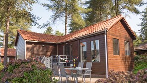 Eine Holzhütte mit Ziegeldach, eine Terrasse mit Stühlen und Tisch, umgeben von Grünpflanzen und Bäumen.