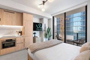 Una habitación de hotel moderna con kitchenette, una cama, un ventanal grande con vista a la ciudad y un cartel que dice 'host'.