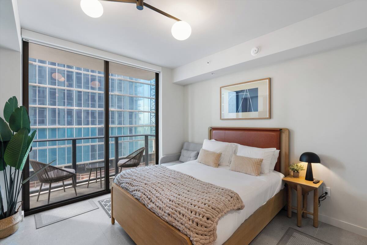 Quarto moderno com uma cama grande, mesa de cabeceira com abajur, quadro na parede e porta de vidro deslizante para uma varanda com cadeiras.