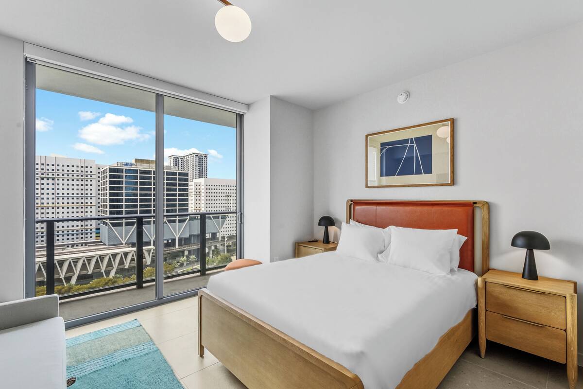 Quarto de hotel moderno com uma cama grande, uma mesinha de cabeceira, um quadro emoldurado e vista da cidade através de janelas do chão ao teto.