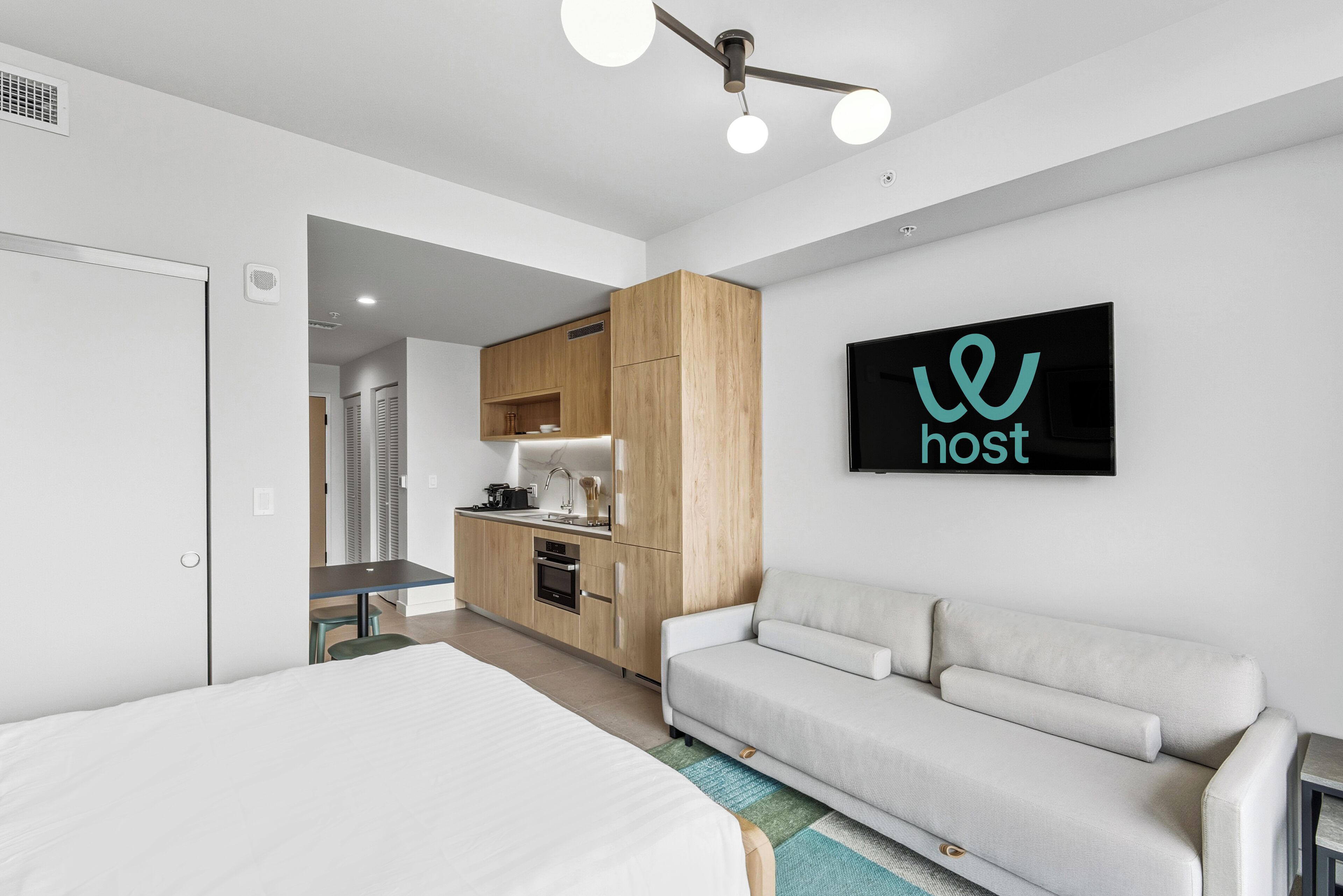 A modern hotel room with a large bed, a sofa, a kitchenette, and a flat-screen TV mounted on the wall.