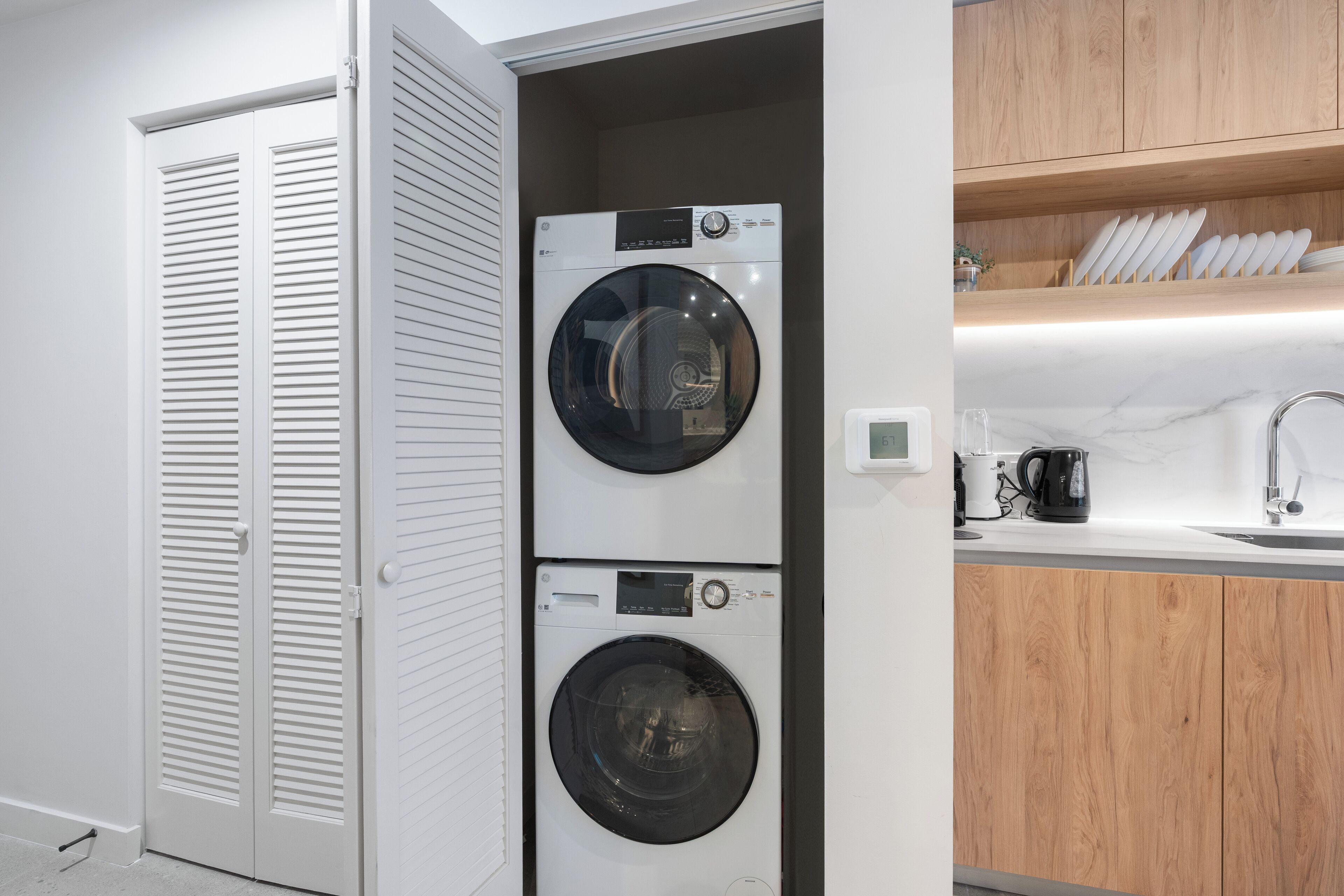 A compact laundry room with two stacked washing machines, a white door with louvered panels, and a wooden countertop with a coffee maker and plates.