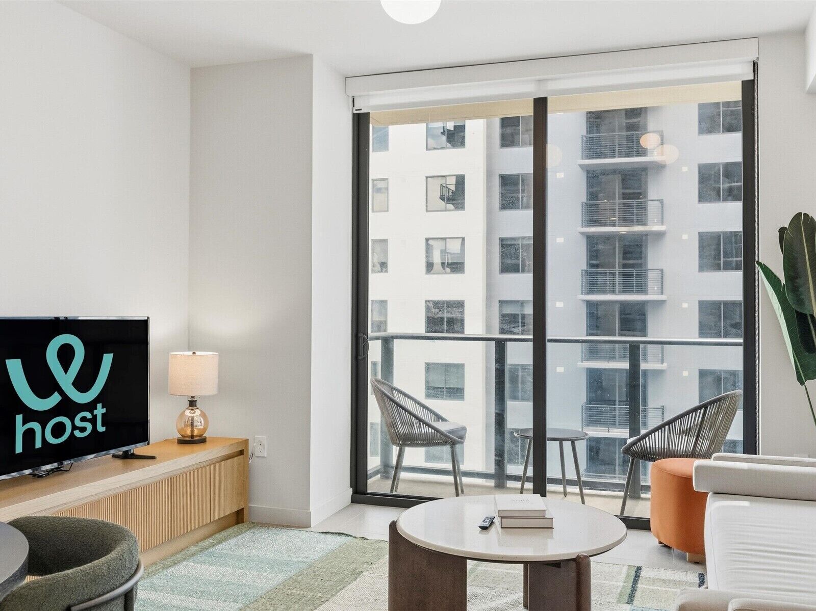 A modern hotel room with a large window, a flat-screen TV, a wooden desk, a lamp, and two chairs.