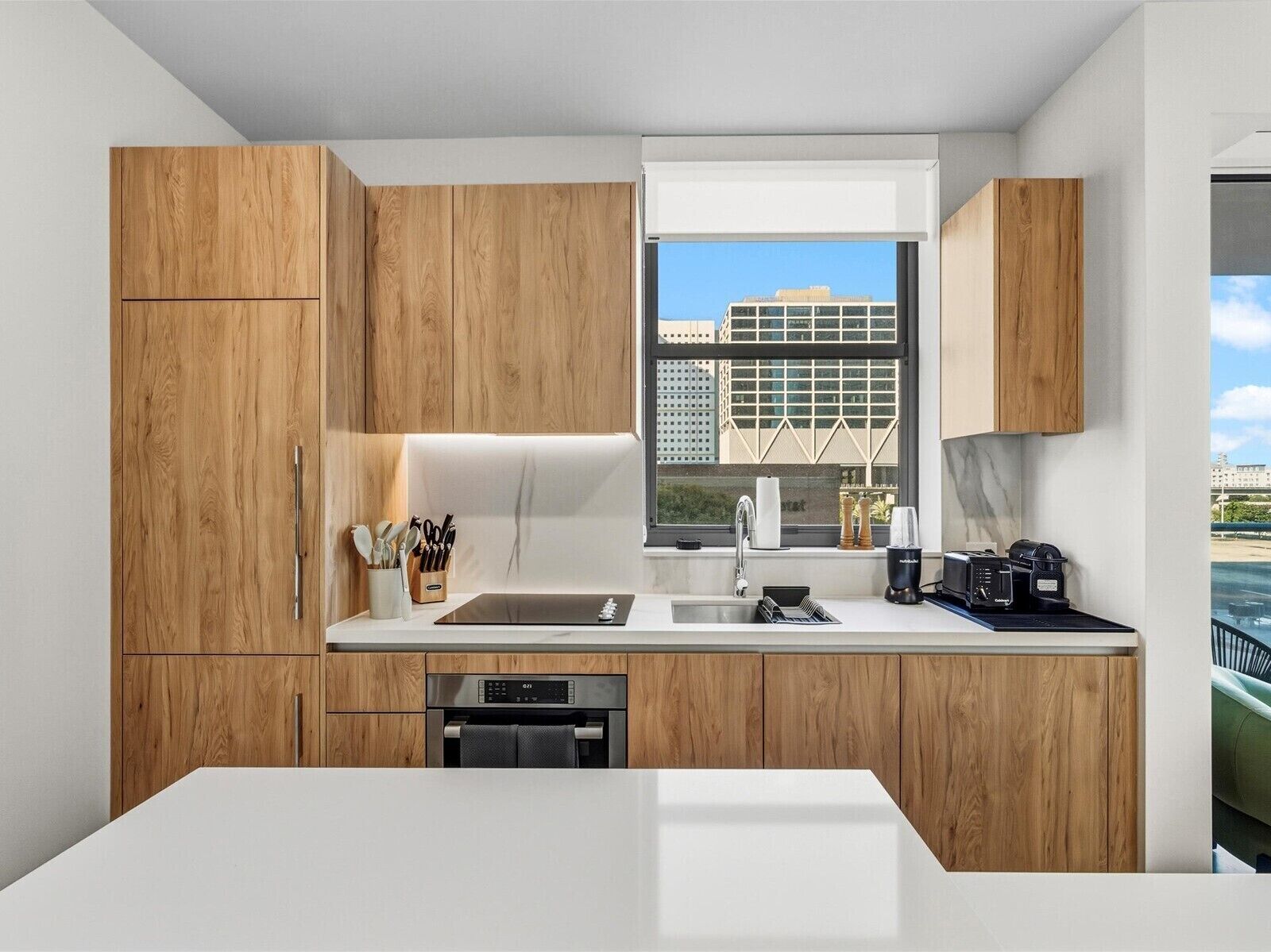 Una cocina moderna con armarios de madera, horno empotrado, lavamanos y ventana con vista a la ciudad.