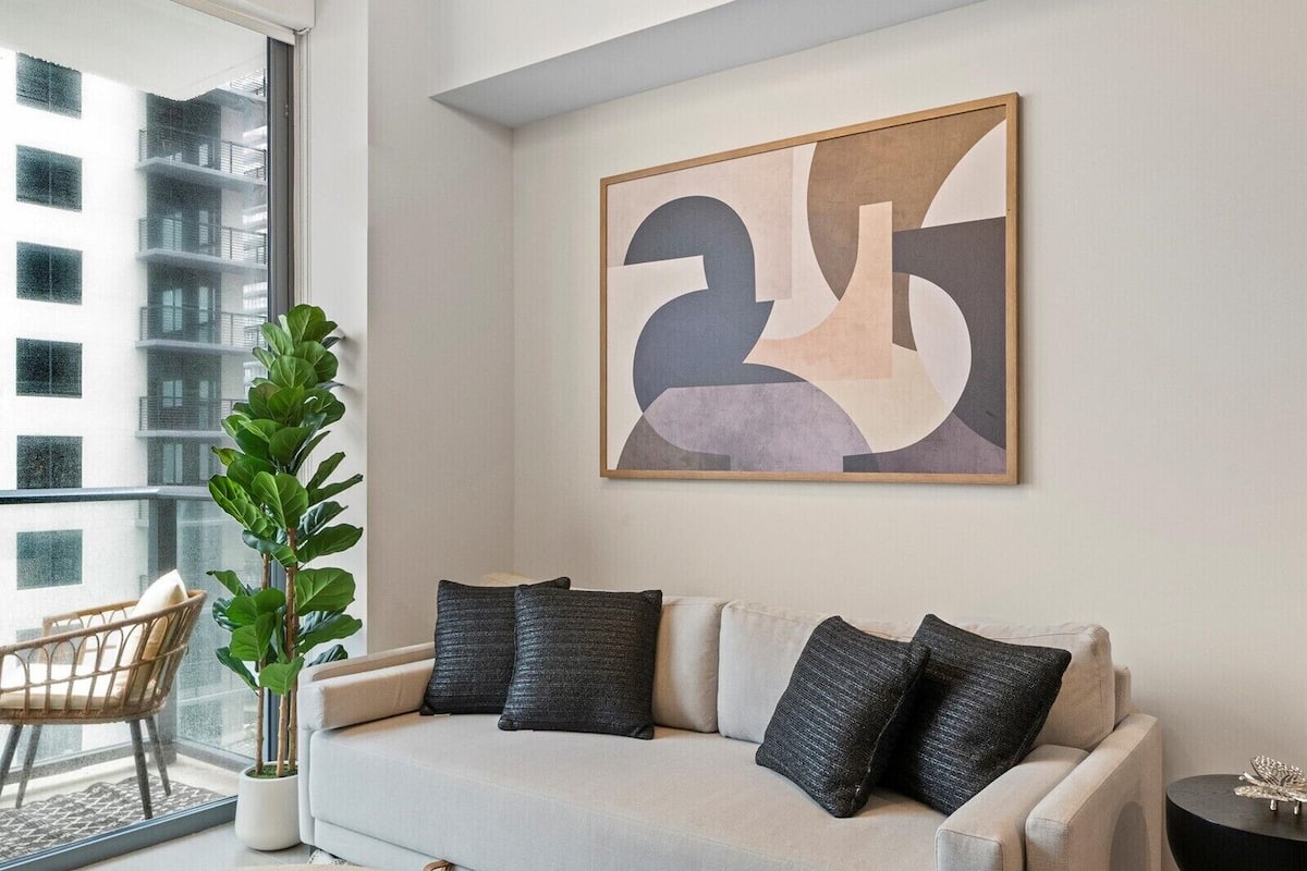 Sala de estar moderna com sofá cinzento, almofadas pretas, um quadro abstrato grande, uma planta em vaso e vista para um edifício pela janela.