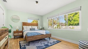 Un dormitorio con cama, mesita de noche, cómoda, ventana con vista a árboles y ventilador de techo.