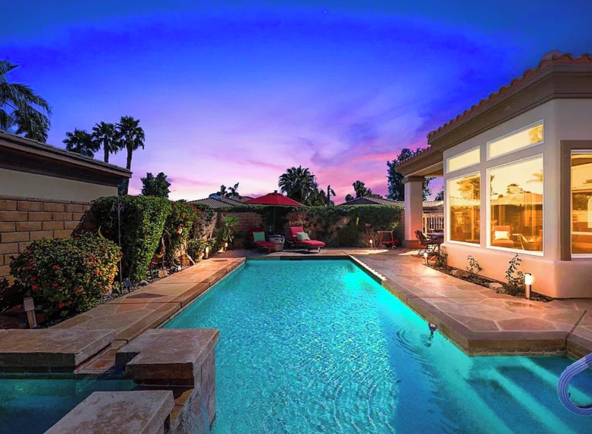 A swimming pool with a patio area, surrounded by a landscaped garden and a building with large windows.