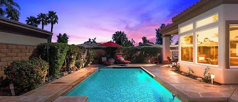 A swimming pool with a patio area, surrounded by a landscaped garden and a building with large windows.