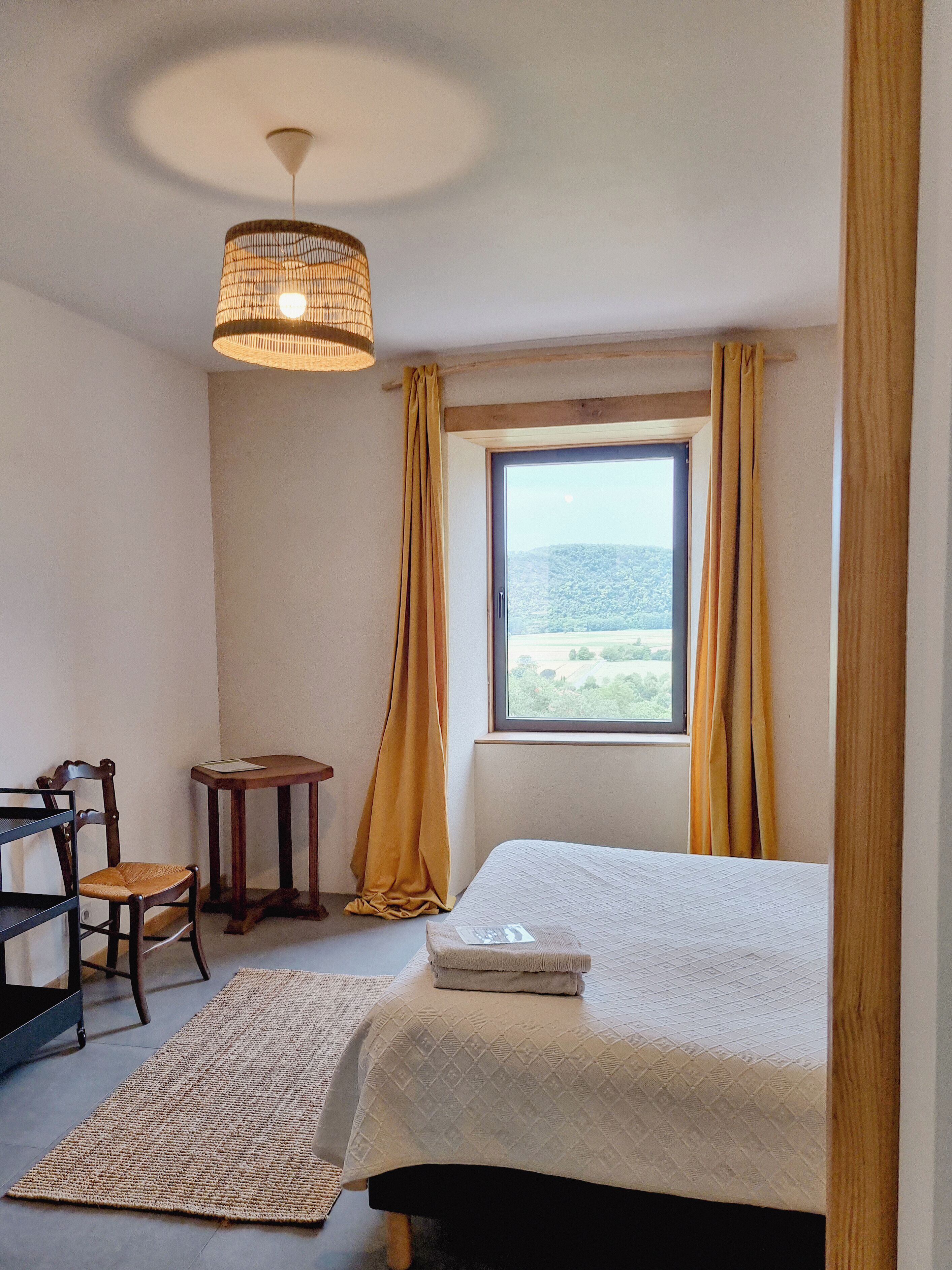 Een slaapkamer met een bed, een stoel, een tafeltje, een raam met gordijnen en een hanglamp.