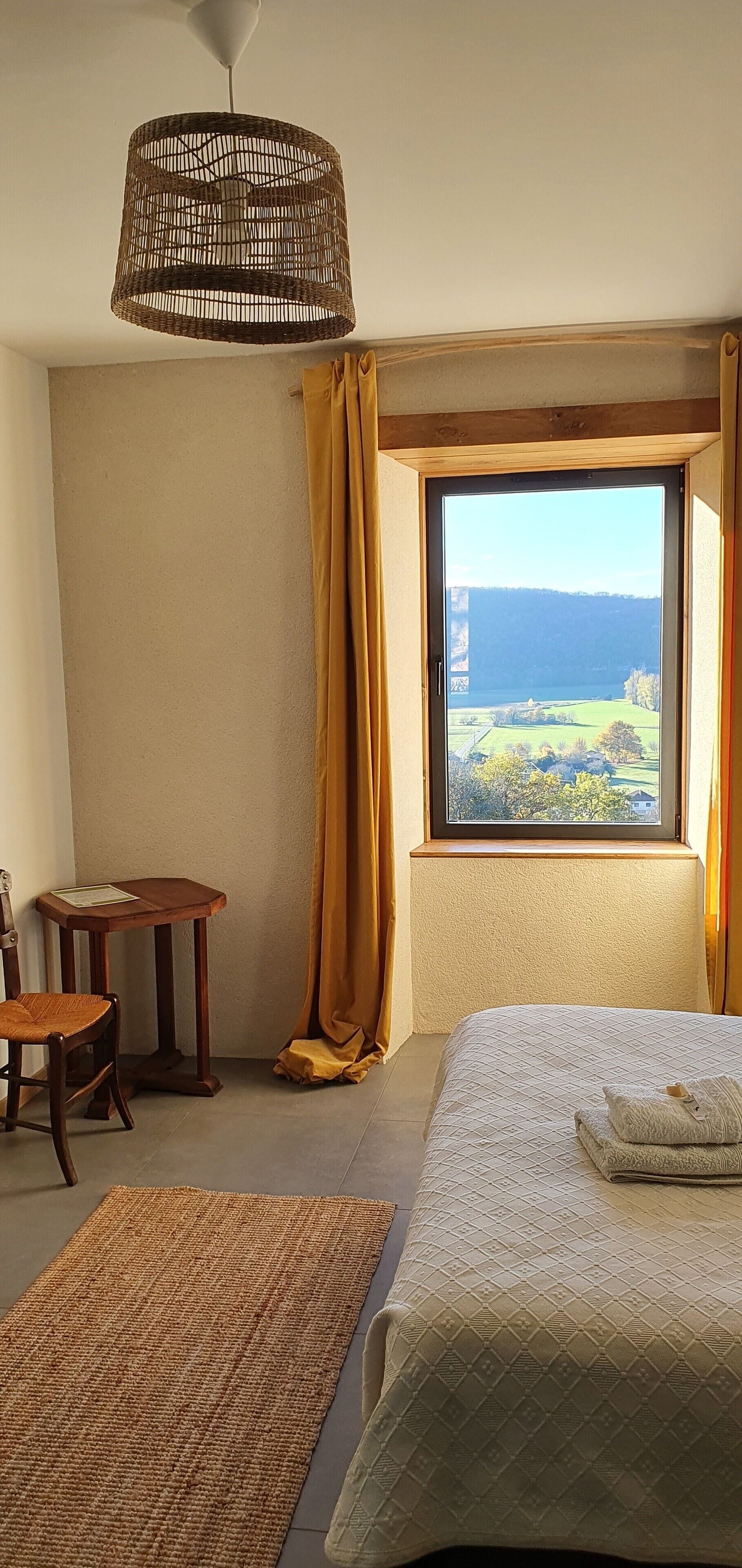 A bedroom with a bed, a small wooden table, a chair, a woven floor lamp, and a large window with a view of a landscape.