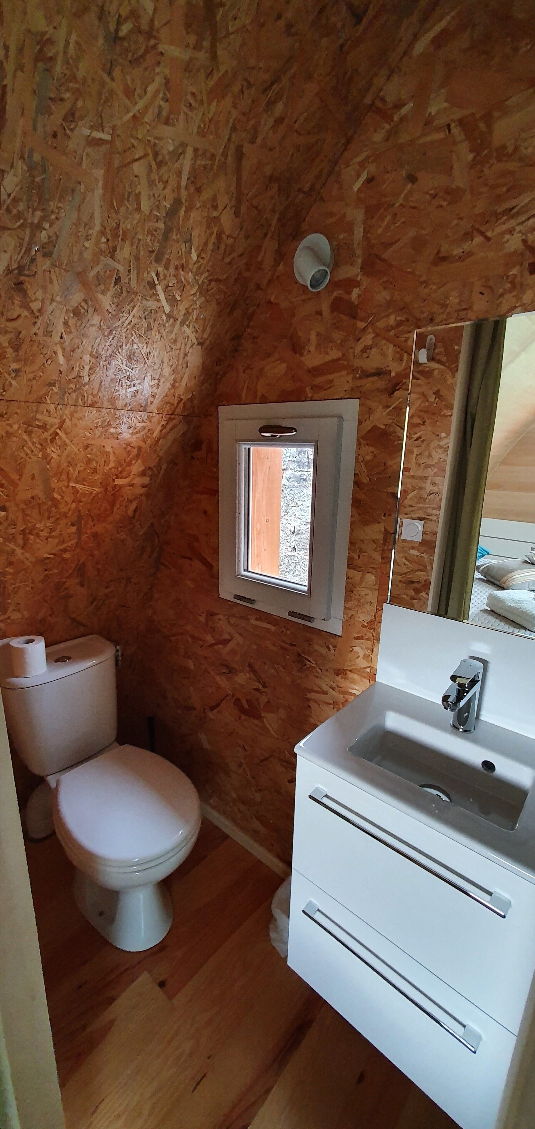 A small, compact bathroom with a wooden interior, a toilet, a sink, and a window providing natural light.