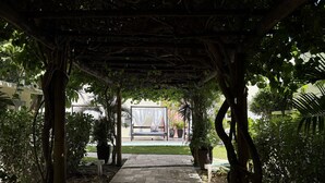 Un paseo techado con un banco, rodeado de abundante vegetación y una vista despejada de un edificio al fondo.