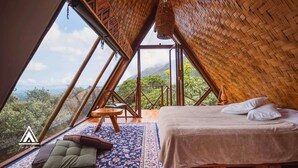 A triangular room with a bed, a small table, and a chair, featuring a large window and a view of the outdoors. - Chalé Tipi da Serra (Brumadinho)