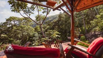 Um deck de madeira com cadeiras de descanso e uma almofada vermelha, com vista para uma floresta exuberante e colinas.