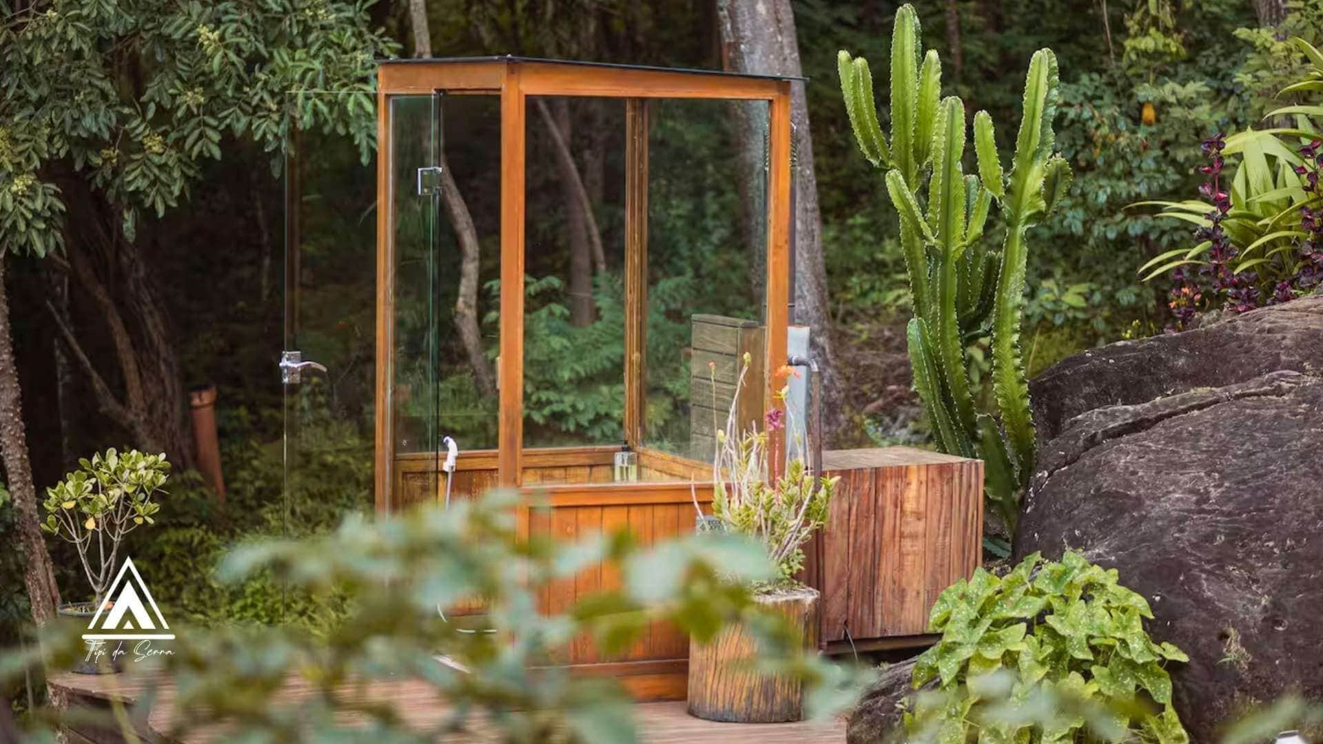 Uma estrutura de madeira com paredes de vidro, contendo um banco de madeira, rodeada por vegetação exuberante e cactos.