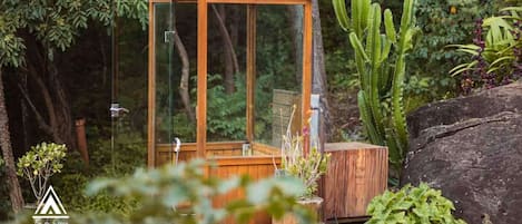 Une structure en bois à l’intérieur de laquelle on trouve un banc en bois, le tout protégé par des parois de verre et entouré d’une végétation luxuriante et de cactus.
