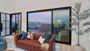 A modern living room with a leather sofa, large windows, and a view of a desert landscape. - The Lookout- Luxe, Hot Tub, Firepit, 50-Mile Views (Page)