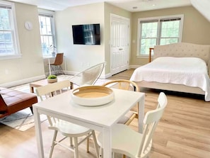 Quarto moderno com cama, televisão, área de refeições com mesa e cadeiras, e uma janela grande.