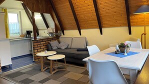 Une pièce chaleureuse avec un plafond en bois, un canapé agrémenté de coussins à motifs, une table à manger avec des chaises, et un coin cuisine doté d’une fenêtre et d’un évier.
