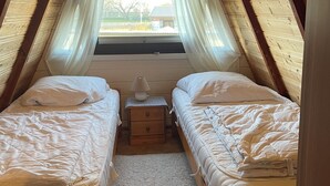 Une petite chambre mansardée avec deux lits simples, une table de chevet, une fenêtre avec des rideaux légers et un plafond en bois.