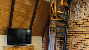 Une pièce chaleureuse avec un plafond en bois, un mur en briques, un téléviseur posé sur un meuble en bois, une chaise blanche et une petite table sur laquelle se trouve un panier rempli de fleurs.