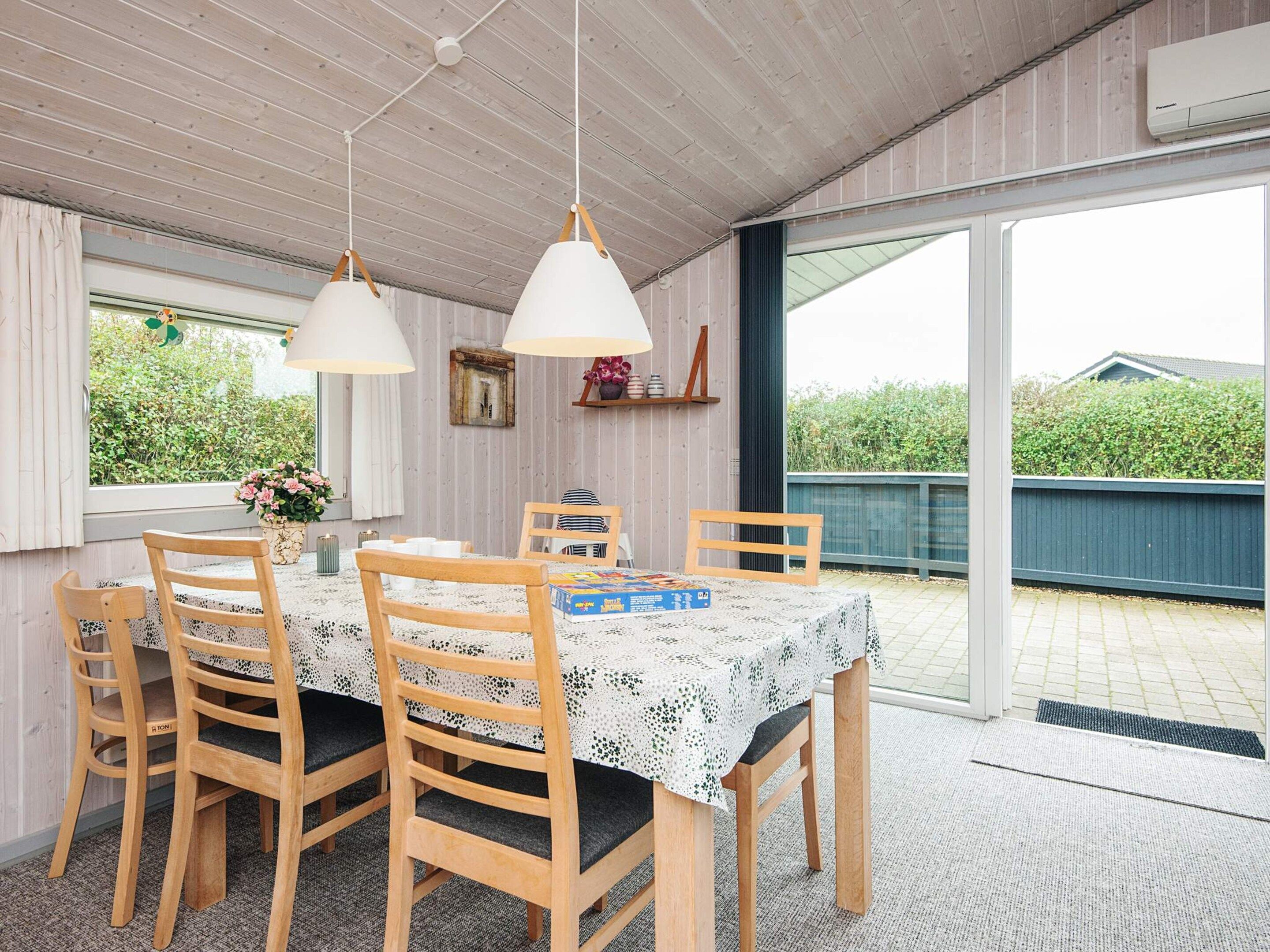 A dining area with a table set for four, wooden chairs, pendant lights, and a view through a glass door.