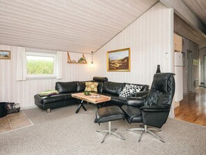 Ein geräumiges Wohnzimmer mit einer schwarzen Ledersofa, einem Holztisch und einem schwarzen Sessel. Zwei Fenster sorgen für natürliches Licht, ein an der Wand befestigtes Regal zeigt Dekorationsobjekte, und über der Couch hängt ein Gemälde.