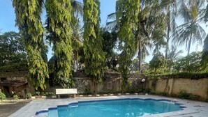 Une piscine dont les bords sont recouverts de carreaux et qui est bordée de briques, entourée de palmiers et d’un mur de pierre.