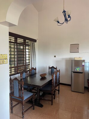 A dining area with a wooden table, chairs, a chandelier, a window with blinds, and a refrigerator. - Nyali 1BR spacious with King Bed, pool, ensuite (Mombasa)