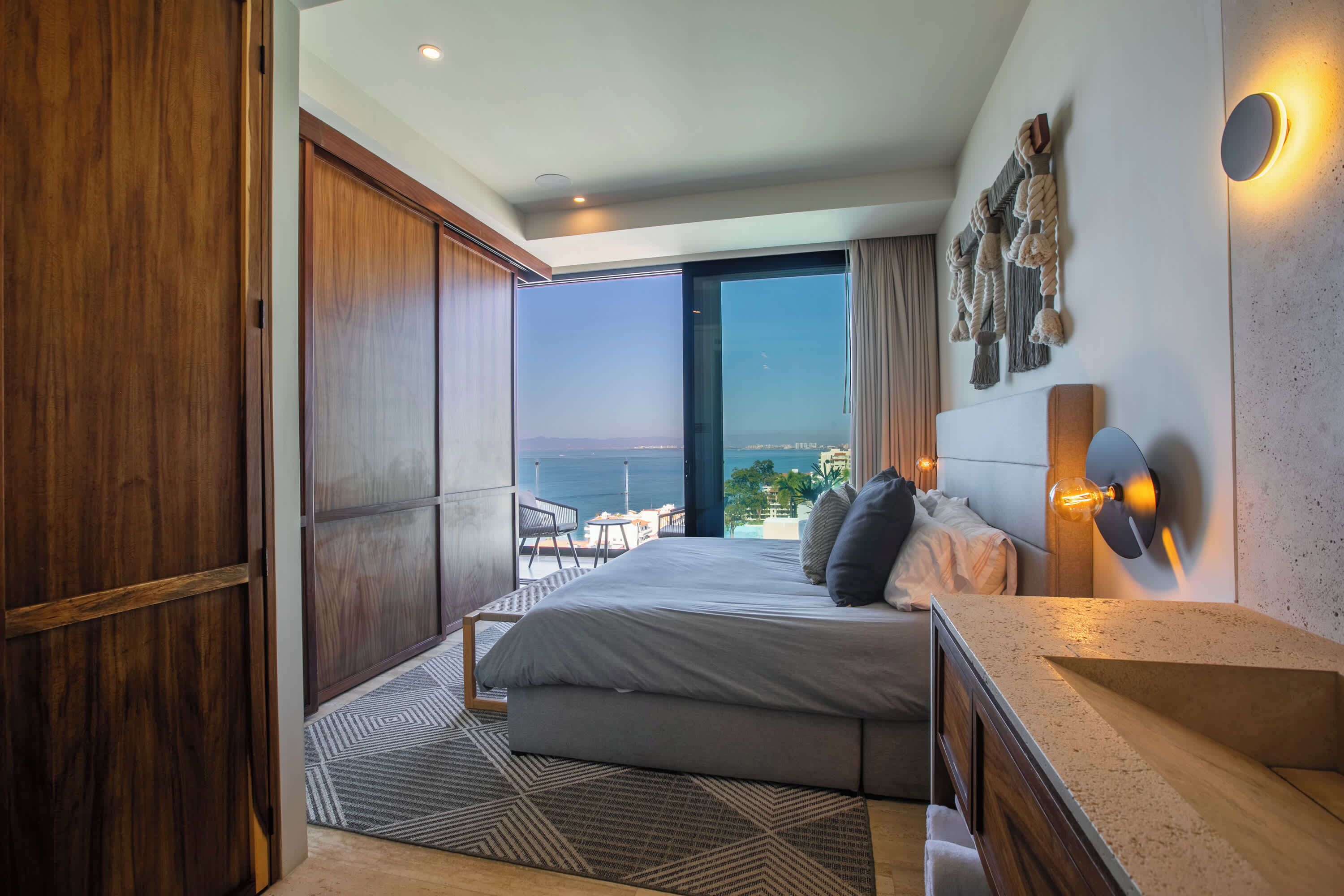 Une chambre moderne avec un grand lit, une vue sur la mer, une armoire en bois et une table de chevet avec une lampe.