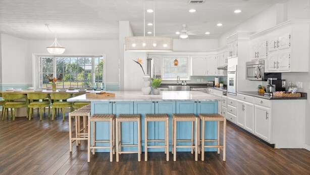 A modern kitchen with a large island, white cabinetry, and a dining area with green chairs. - Sleeps 20! Pickleball, Heated Pool, Hot Tub & Games Retreat! Near Beaches (Bradenton)