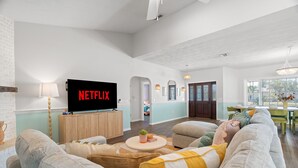 A spacious living room with a large flat-screen TV displaying 'NETFLIX', a comfortable sofa set, a small round coffee table, and a dining area with a green table and chairs. - Sleeps 20! Pickleball, Heated Pool, Hot Tub & Games Retreat! Near Beaches (Bradenton)